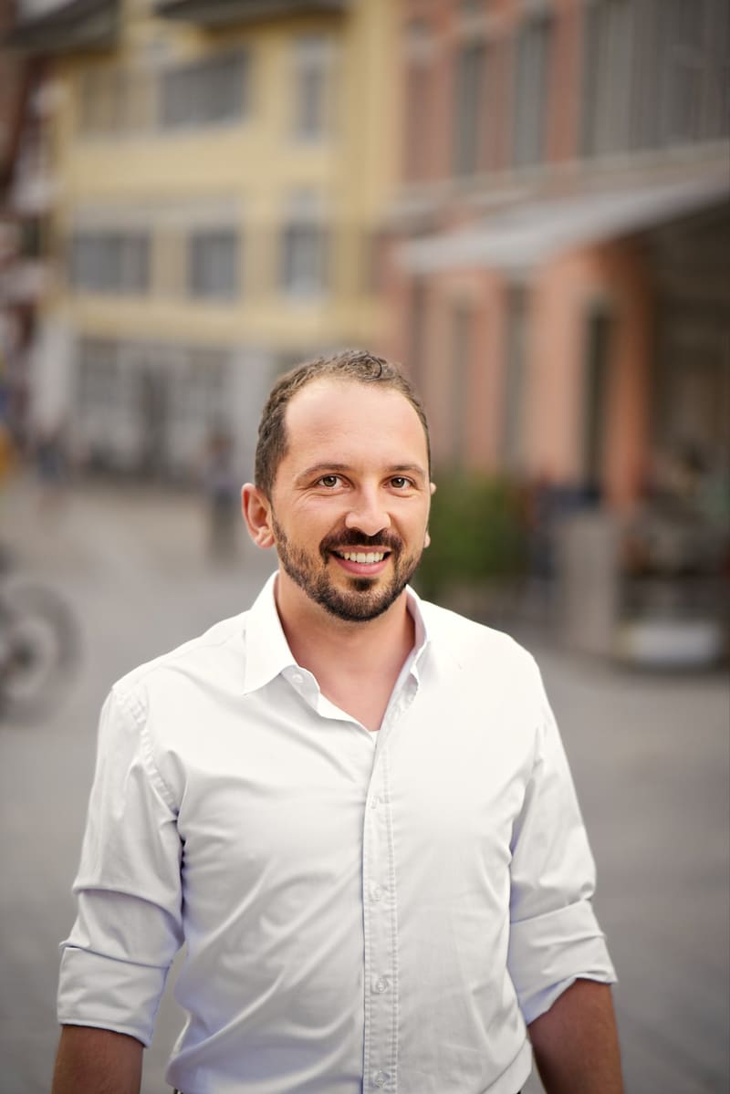 Porträt von Lukas Affolter im weissen Hemd, er lächelt, im Hintergrund unscharf eine Gasse.